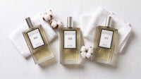 Three TEV perfume bottles - No. 821, No. 452, No. 111 - on white linen with cotton blossoms and soft morning light